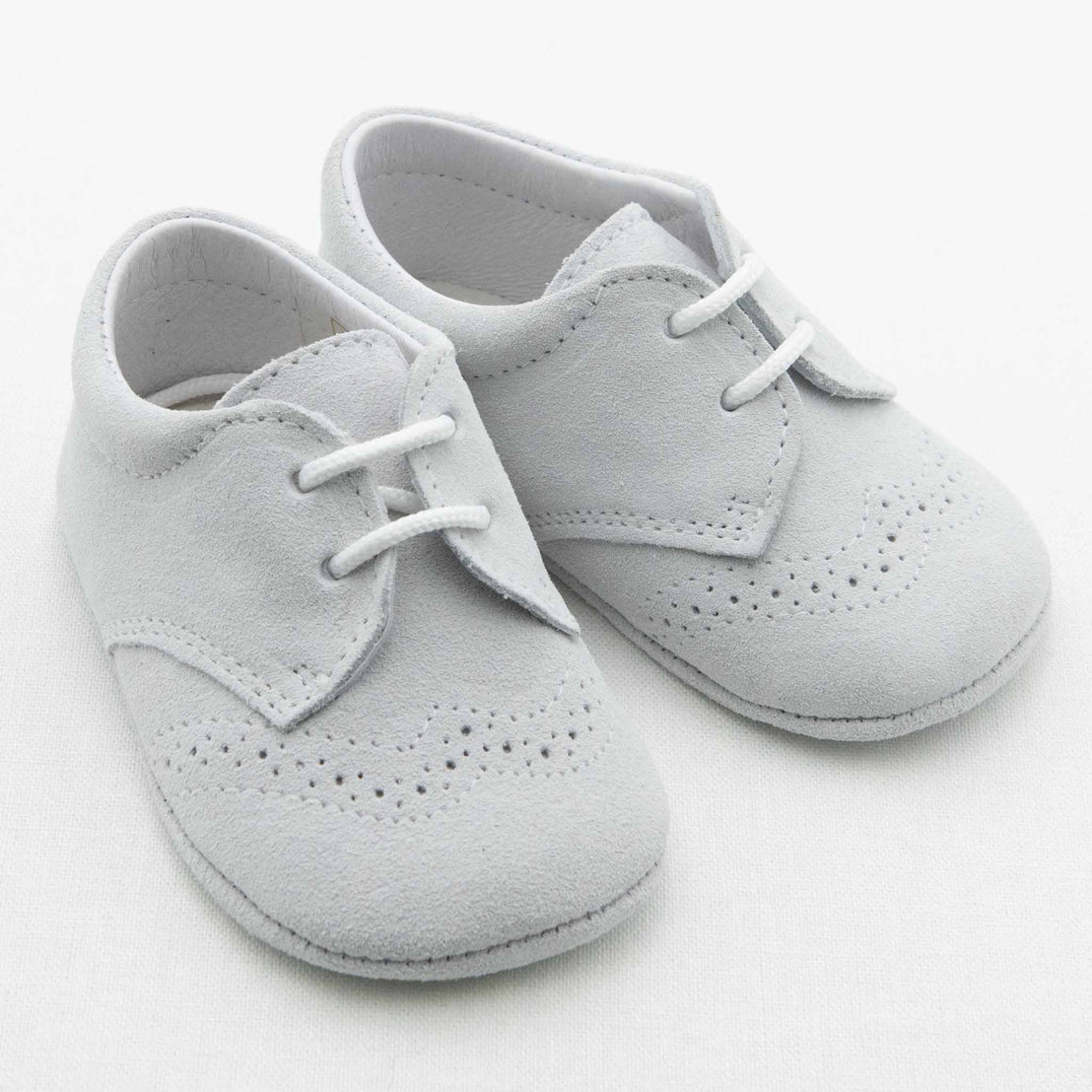 A pair of Miles Suede Shoes by Baby Beau & Belle, featuring dove grey suede with decorative perforations on the toes and sides. Handmade in Spain, these infant shoes have white laces and soft soles, ideal for babies. The background is a plain white surface.