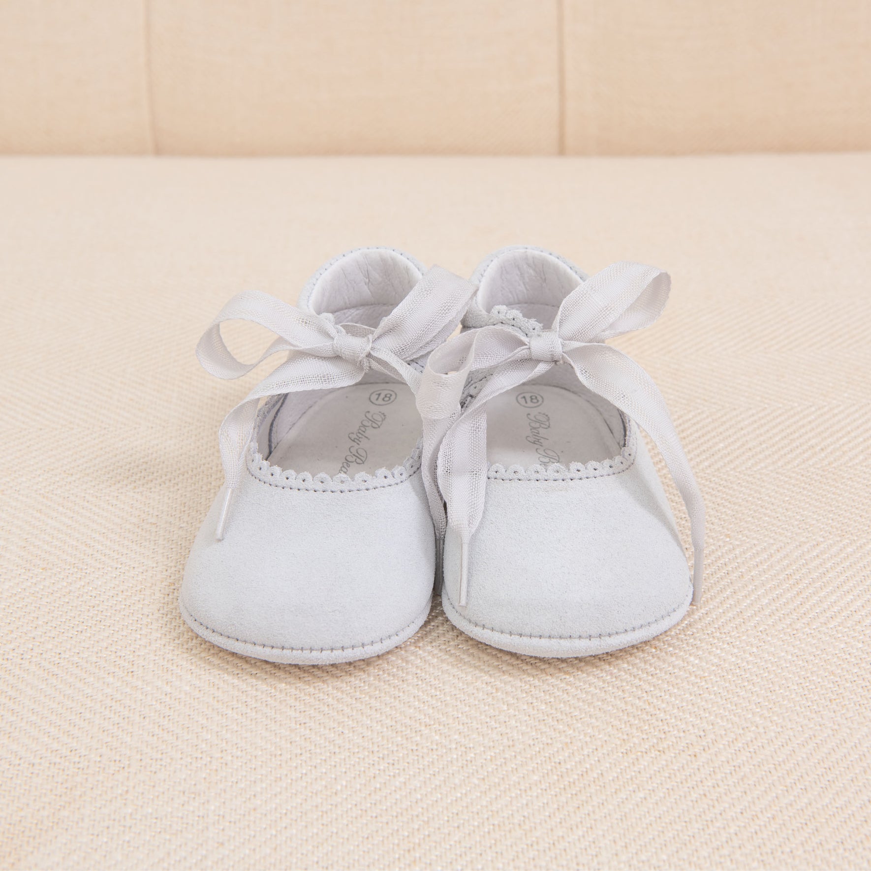 A pair of Dove Grey Suede Tie Mary Janes, handmade in Spain from suede leather with ribbon ties, is placed on a beige fabric surface. The shoes have a decorative edge near the opening and are neatly arranged with their ribbons tied in bows.