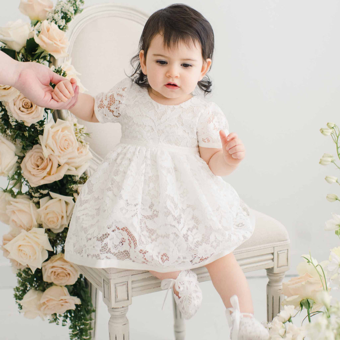 A toddler with dark hair in a white Rose Romper Dress sits on a cushioned stool, holding an adult's hand, surrounded by cream roses on a white background.