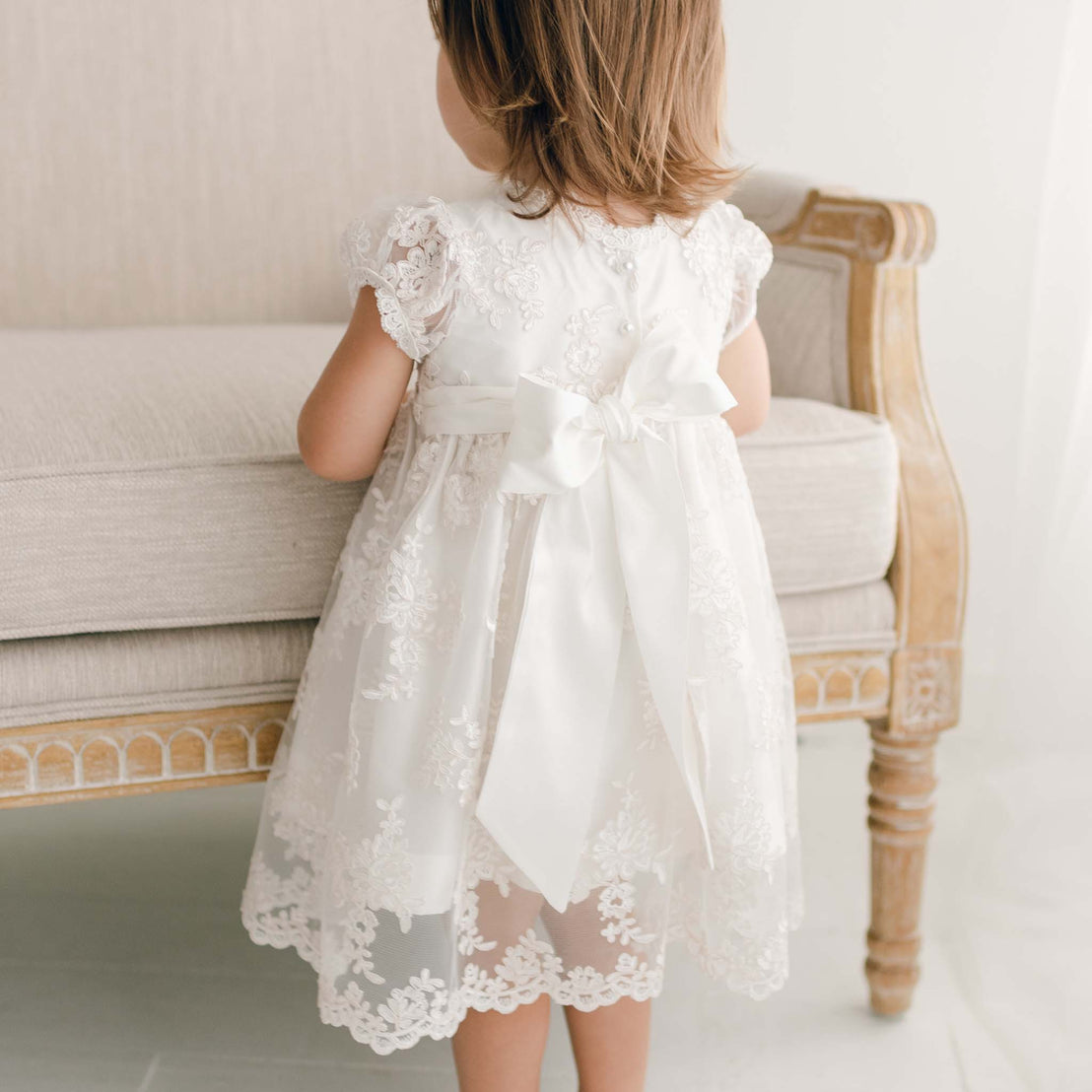 Back detail of Penelope christening dress. Young toddler standing in baptism outfit next to chair.