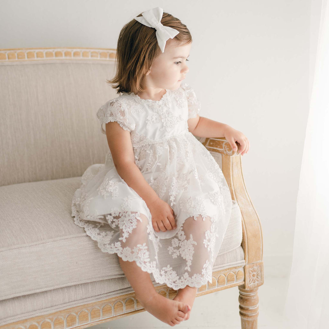 Photo of young toddler sitting on couch wearing her lace christening dress, part of the Penelope baptism collection.