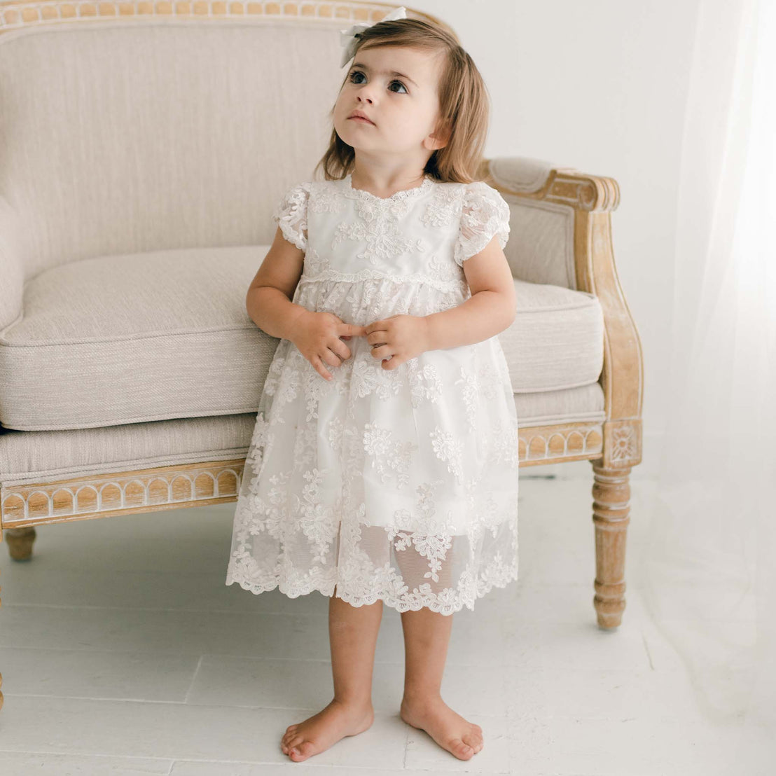 Young toddler standing by couch wearing the Penelope lace christening dress & bloomers.