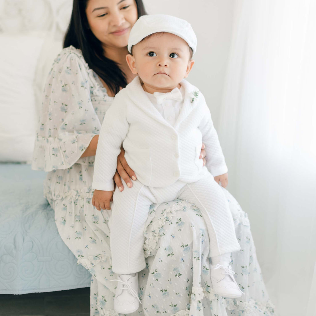 A young child wearing the Elijah 3-Piece Set, which includes a white christening suit and matching cap, along with white shoes, sits on the lap of a woman in a light floral dress. They are indoors in a well-lit room. The woman has long dark hair and is smiling while looking at the child.