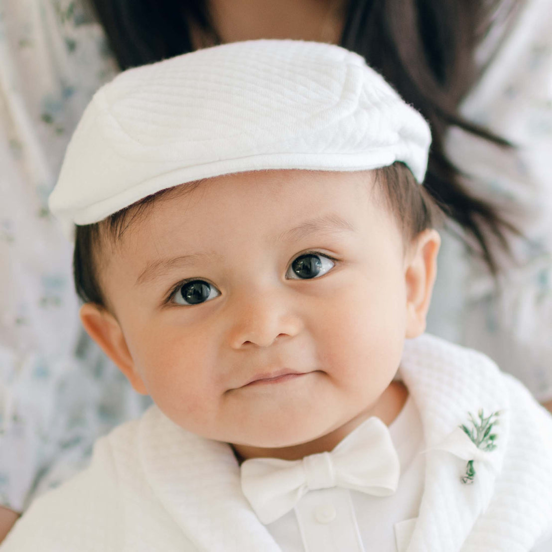 A baby dressed in white textured cotton, wearing the Elijah Newsboy Cap along with a matching bow tie, looks towards the camera. The baby has dark eyes and a small smile. Partial view of an adult in the background.