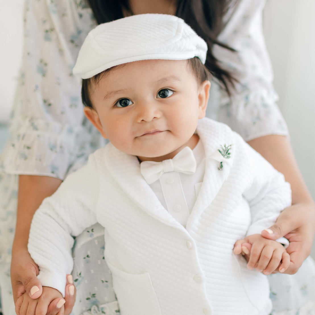 A baby boy dressed in the Elijah 3-Piece Set, which includes a white christening suit with a bow tie and cap, is standing while holding the hands of an adult behind him. The baby is looking at the camera with a neutral expression. The adult's body is partially visible, dressed in a light, textured cotton garment.