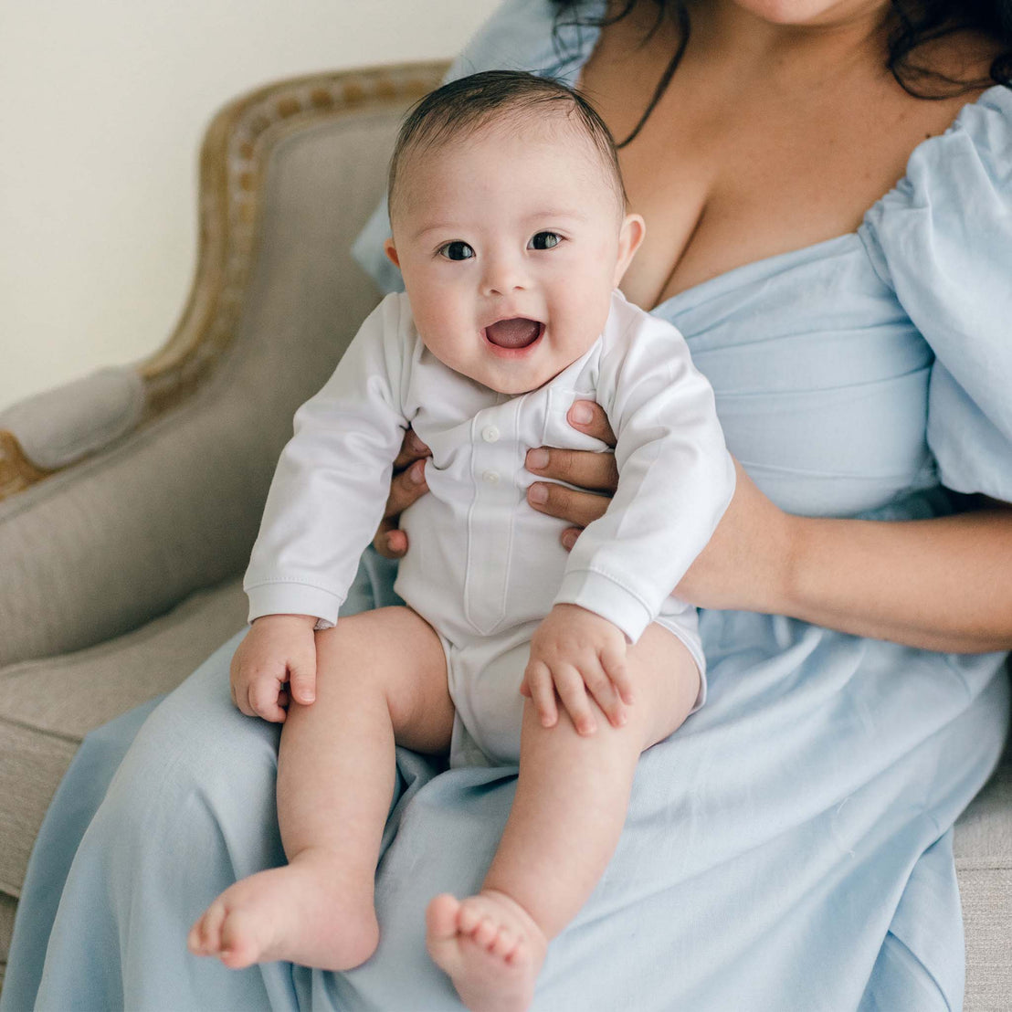 Baby boy sitting on his mother's lap and wearing a white onesie, included in the Elijah 3-Piece Suit, and made from 100% cotton adorned with a faux placket and buttons.