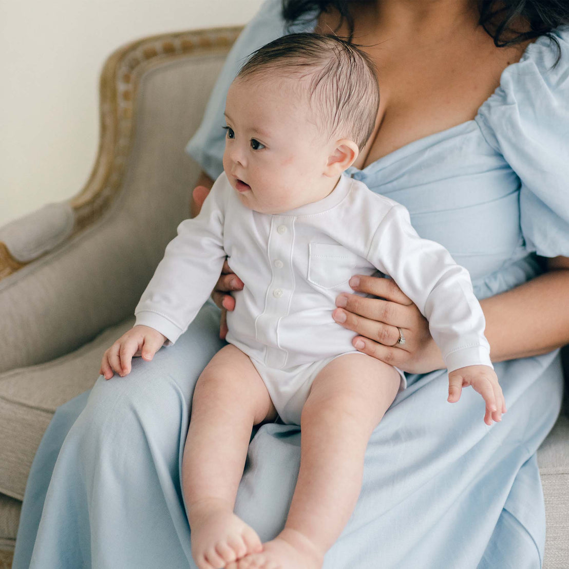 Baby boy sitting on his mother's lap and wearing a white onesie, included in the Elijah 3-Piece Suit, and made from 100% cotton adorned with a faux placket, buttons, and a chest pocket.