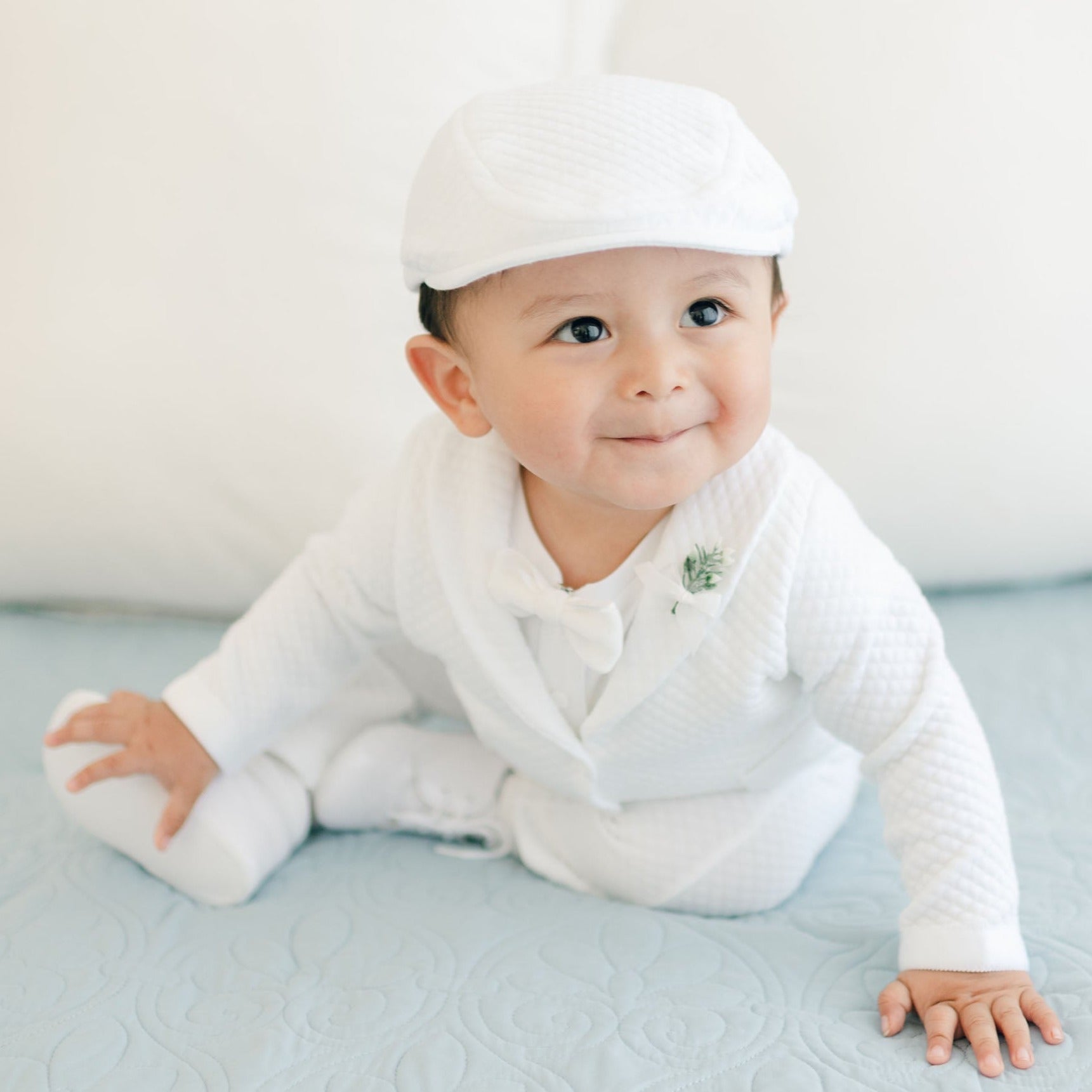 A baby dressed in the Elijah 3-Piece Set, featuring a white christening suit paired with a matching hat, is sitting on a light blue quilt. The baby is looking slightly to the side with a small smile on their face, and white pillows can be seen in the background.