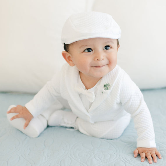A baby dressed in the Elijah 3-Piece Set, featuring a white christening suit paired with a matching hat, is sitting on a light blue quilt. The baby is looking slightly to the side with a small smile on their face, and white pillows can be seen in the background.