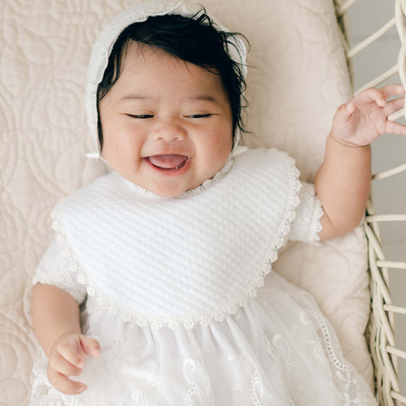 A baby lies in a white crib while wearing a baptism outfit, complete with a lace bonnet and Eliza Bib. The baby is smiling with eyes nearly closed, one hand grasping the side of the crib. The crib sheet has a quilted pattern.