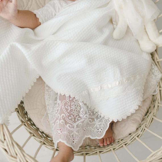 An infant is lying in a wicker basket covered with an Eliza Personalized Blanket. The infant is wearing a white floral lace dress and has a stuffed toy beside them. Only the lower half of the infant's body is visible, with feet peeking out from the dress.