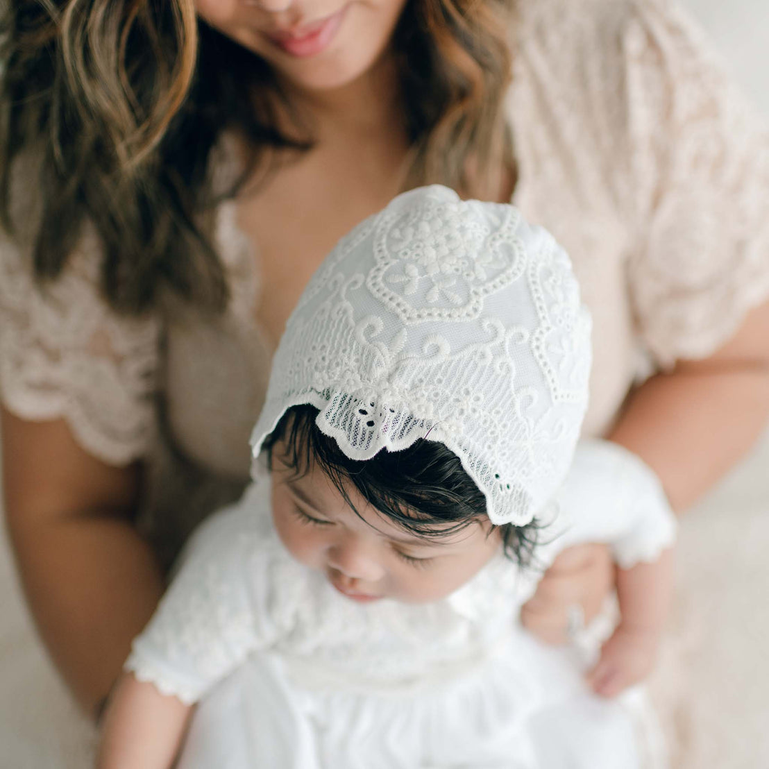 A woman holding a baby dressed in an Eliza Blessing Gown & Bonnet made from 100% pima cotton and adorned with christening accessories. The woman is in soft focus, while the baby takes center stage, gazing downward. The background is neutral, and both are wearing light-colored clothing.