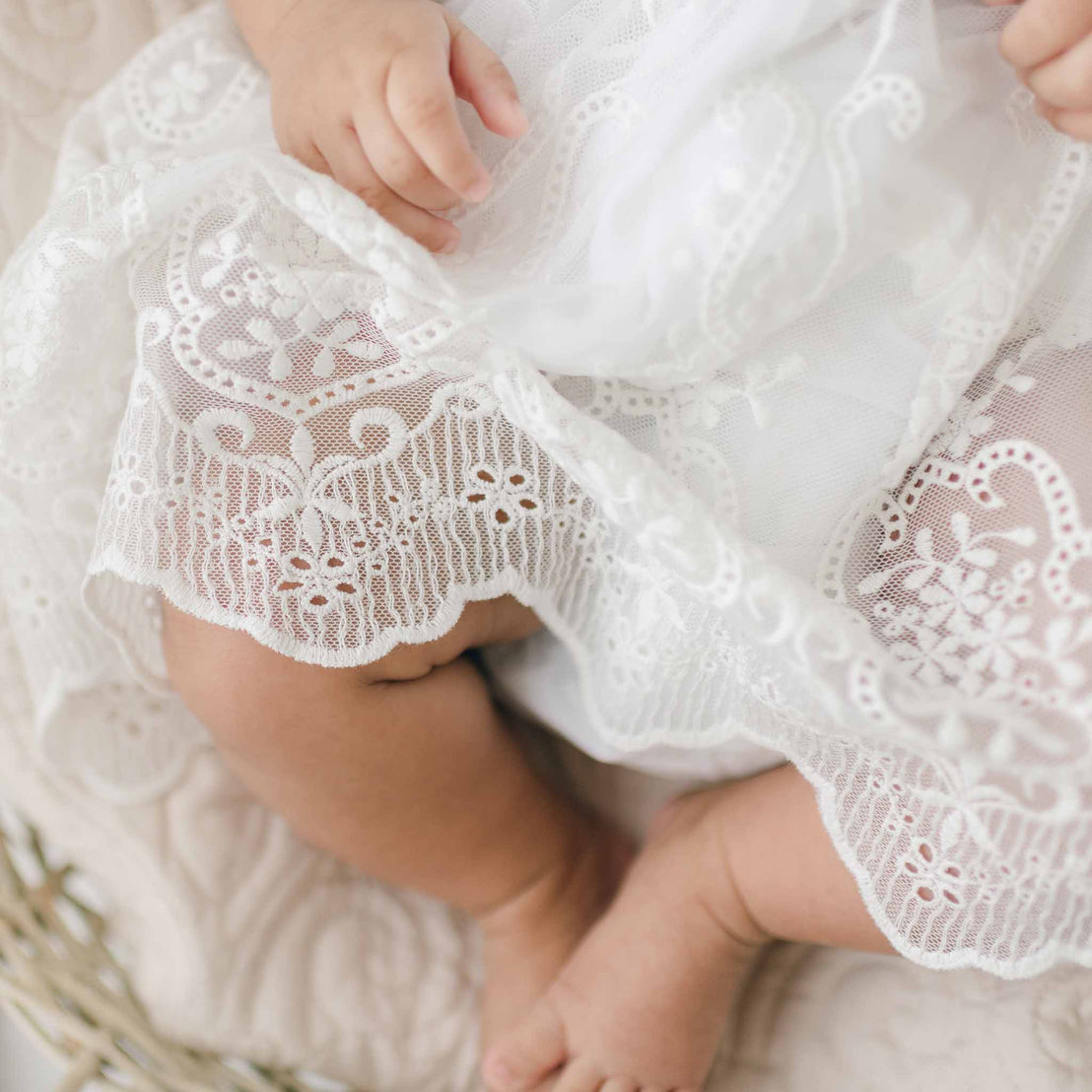 A close-up image of a baby's legs and hands, wearing a delicate Eliza Blessing Dress & Headband made from soft Pima Cotton with intricate floral lace patterns. The baby, seated on a light-colored cushioned surface, showcases the luxurious handmade garment crafted in the USA. Only the lower part of the body is visible.