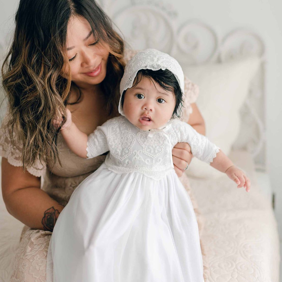 A woman sits on a bed with a baby on her lap. The woman has wavy, shoulder-length hair and is smiling. The baby, dressed in an Eliza Blessing Gown & Bonnet made from 100% pima cotton and adorned with christening accessories, looks at the camera with an open mouth. The background is soft and neutral-toned.