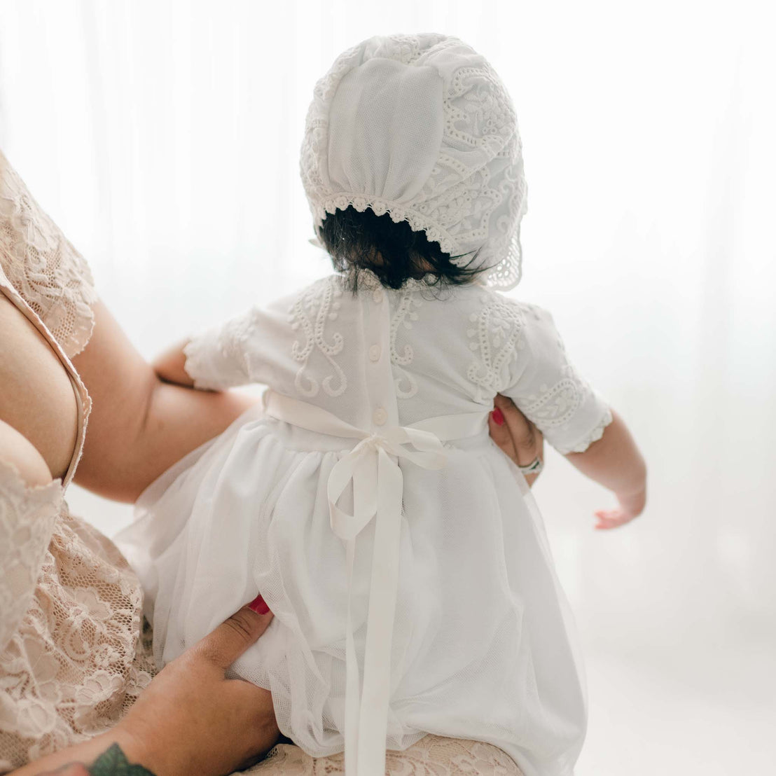 A baby wearing an Eliza Blessing Gown & Bonnet is held by an adult. The baby is facing away from the camera while the adult supports them. The background is softly lit, giving the scene a serene atmosphere, perfect for showcasing Christening accessories.