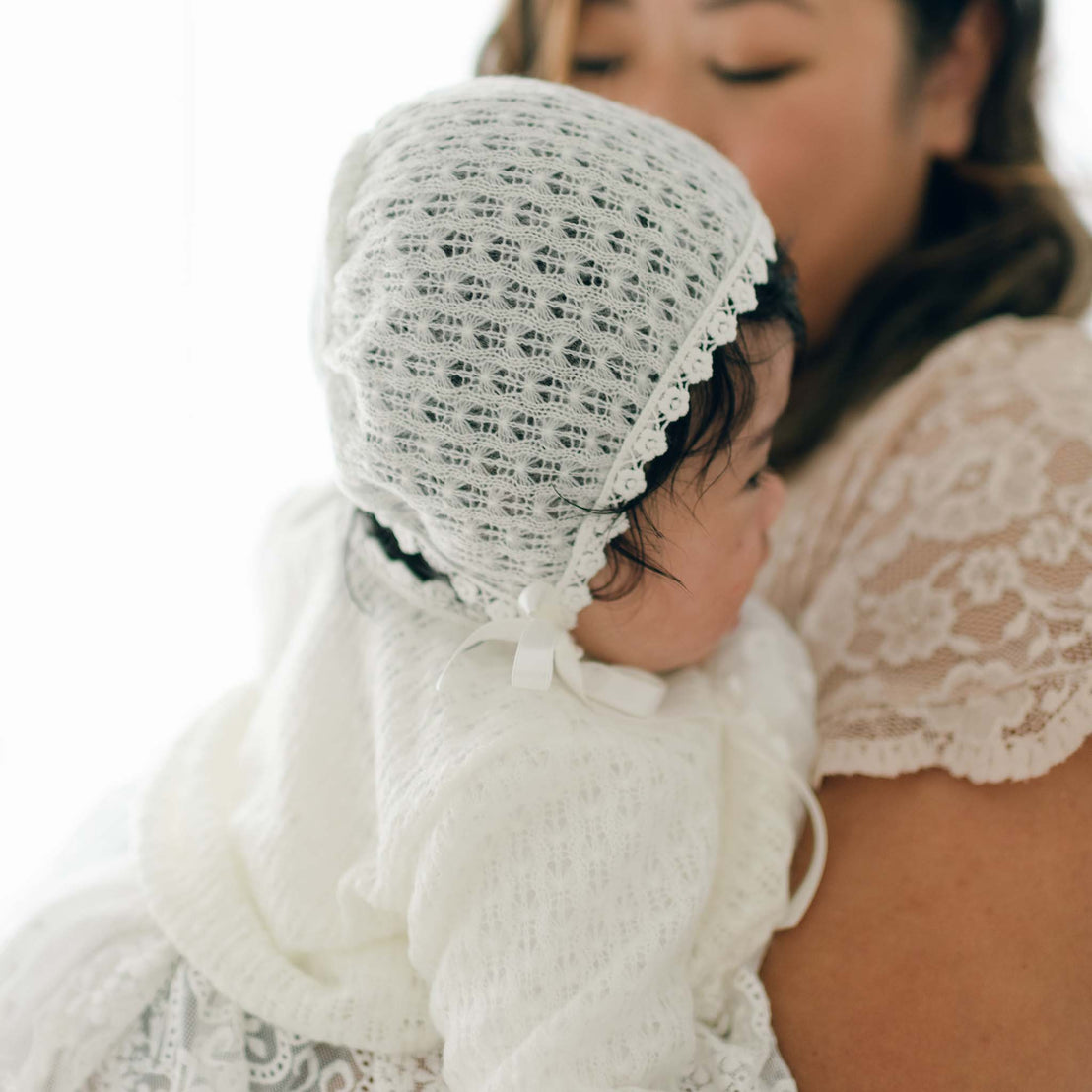 A woman holds a baby dressed in a white lace outfit with silk ribbon ties and an Eliza Christening Knit Bonnet. The woman's face is only partially visible, and she appears to be wearing a lace dress. The image focuses on the baby and the intricate details of their clothing.