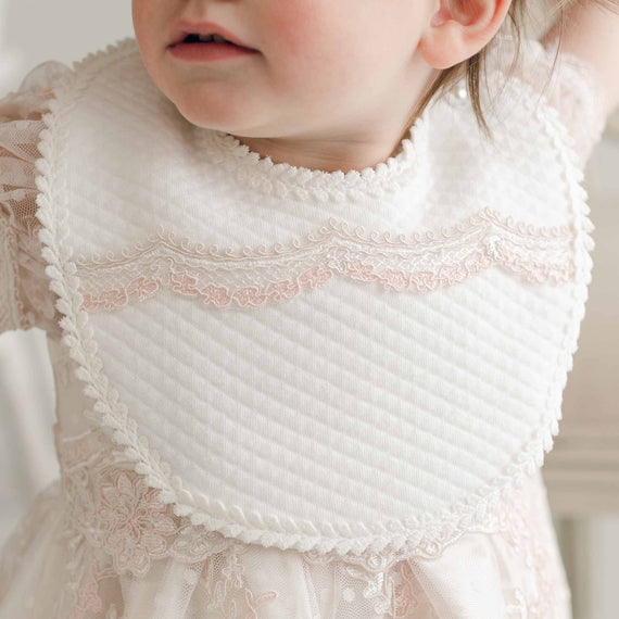 A young child is wearing the Elizabeth Bib, featuring delicate pink lace details. The child is also dressed in a special occasion dress with sheer lace sleeves, looking slightly to the side. The focus is on the upper part of the child's outfit, capturing its vintage charm.
