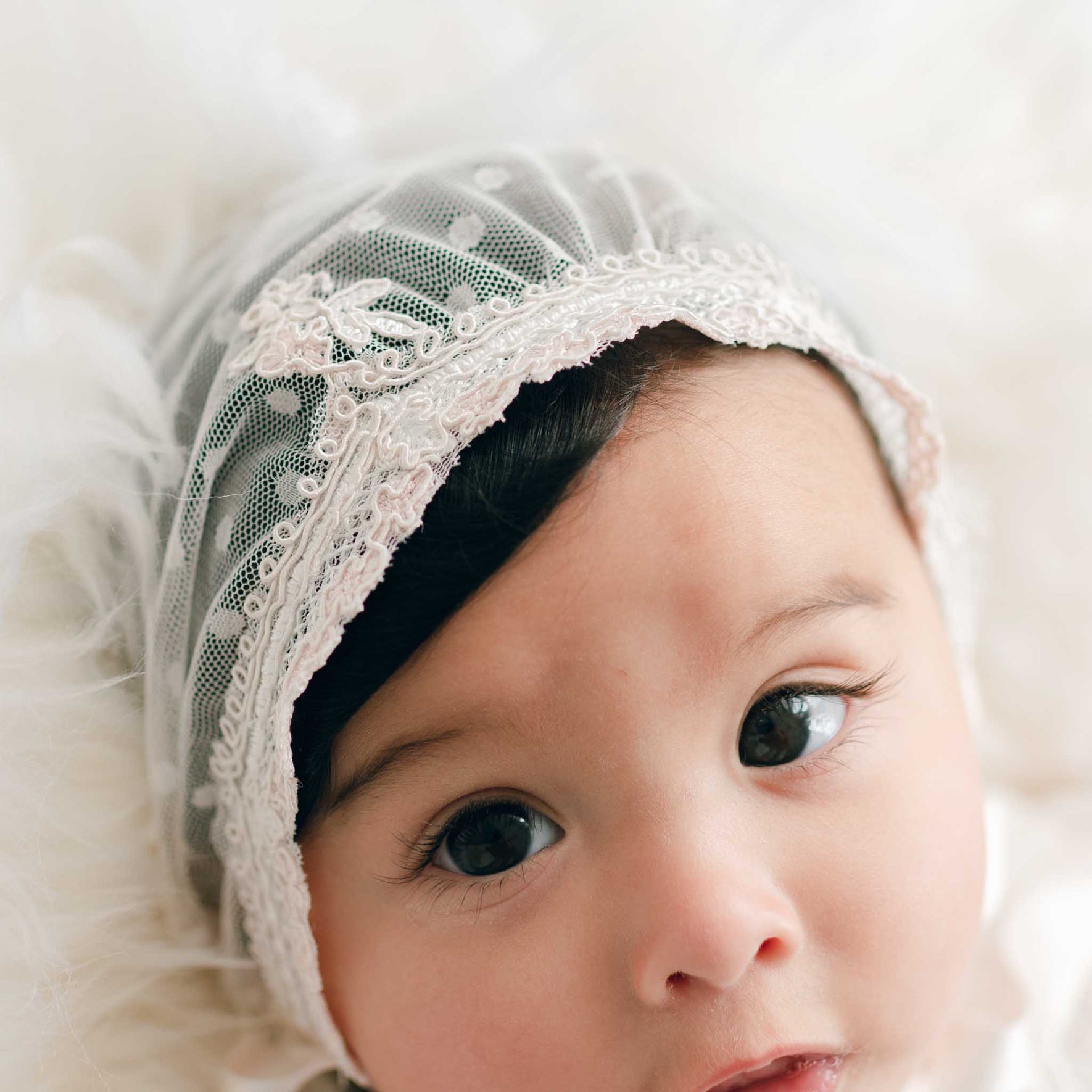 Close-up of a baby wearing a Elizabeth Newborn Christening Bonnet against a soft white background. Details showcase the sheer lace bonnet with silk ribbon ties fit securely around her face, luxurious details characteristic to high end baby clothes.