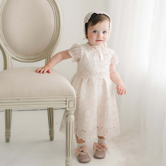 A young child stands next to a white chair in a softly lit room, wearing the exquisite Elizabeth Christening Dress and matching bonnet, paired with pink shoes adorned with bows. One hand rests on the chair while the other hangs by their side. The delicate Christening dress enhances the scene, accompanied by light curtains in the background.