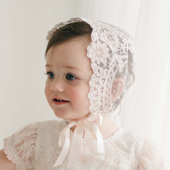 A young child with rosy cheeks is wearing an Elizabeth Lace Bonnet tied with a silk ribbon under the chin. The child is dressed in an ivory and pink lace dress, looking slightly off to the side and smiling. The background is softly lit with white curtains.