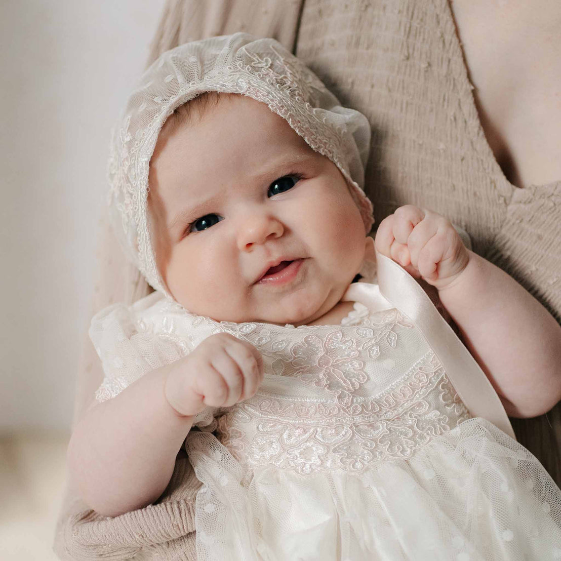 Close-up of a newborn in the Elizabeth Newborn Christening Bonnet and matching Christening Dress held by her mother. Close-up features the details of the delicate lace on the bonnet and dress. Part of the Baby Beau & Belle Baptism Gown Girl Collection.