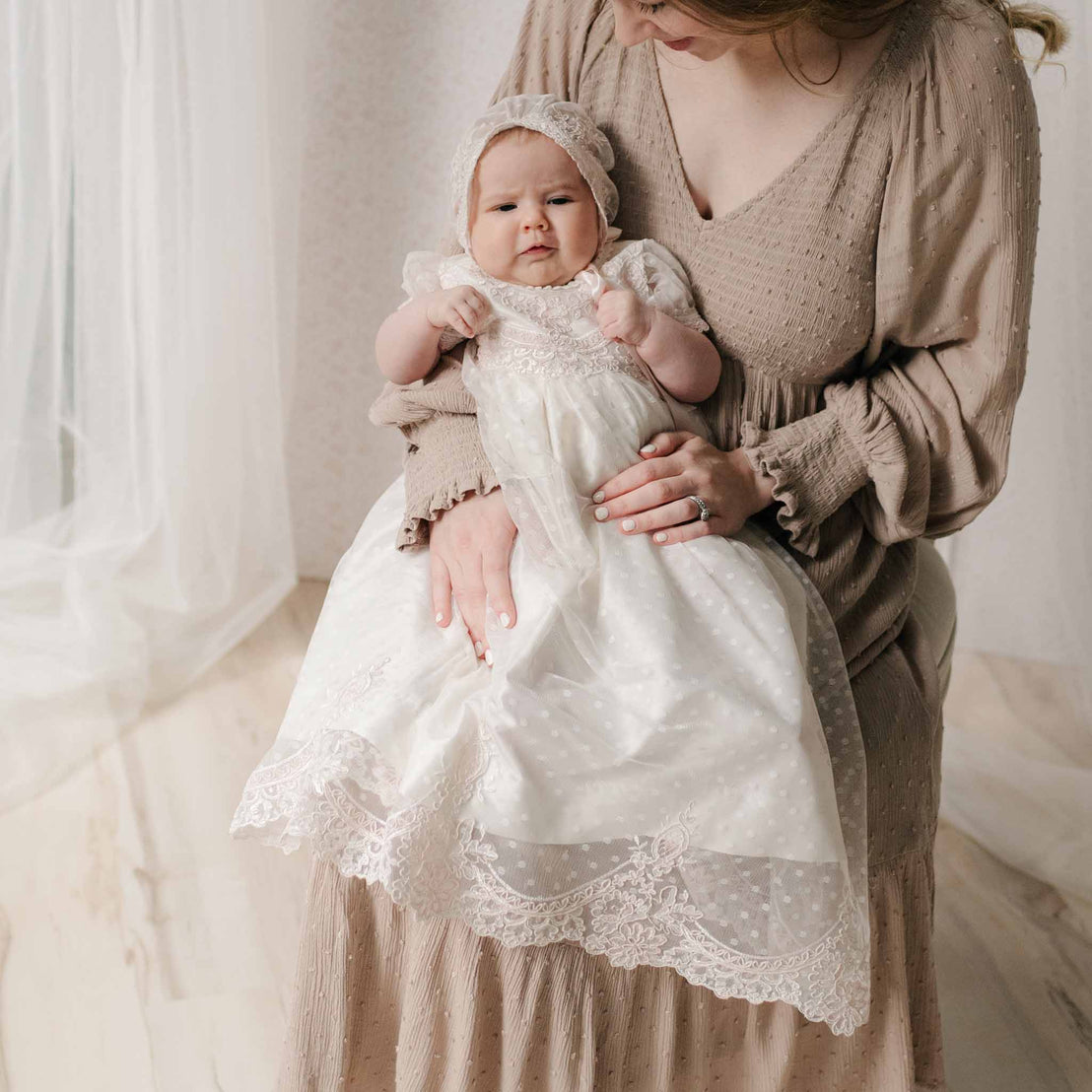 Mother in a beige dress holding a newborn dressed in the Elizabeth Newborn Christening Gown & Bonnet set against a neutral background with a sheer curtain behind. Part of the Baby Beau & Belle Baby Girl Baptism Dress Collection.