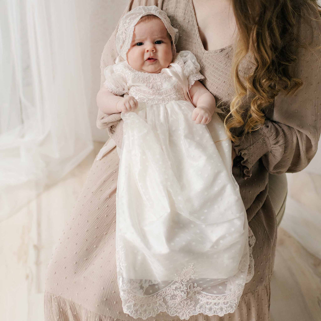 Newborn baby in the Elizabeth Newborn Christening Gown & Bonnet held by her mother in a beige dress against a neutral background. Part of the Baby Beau & Belle Baptism Gown Girl Collection.