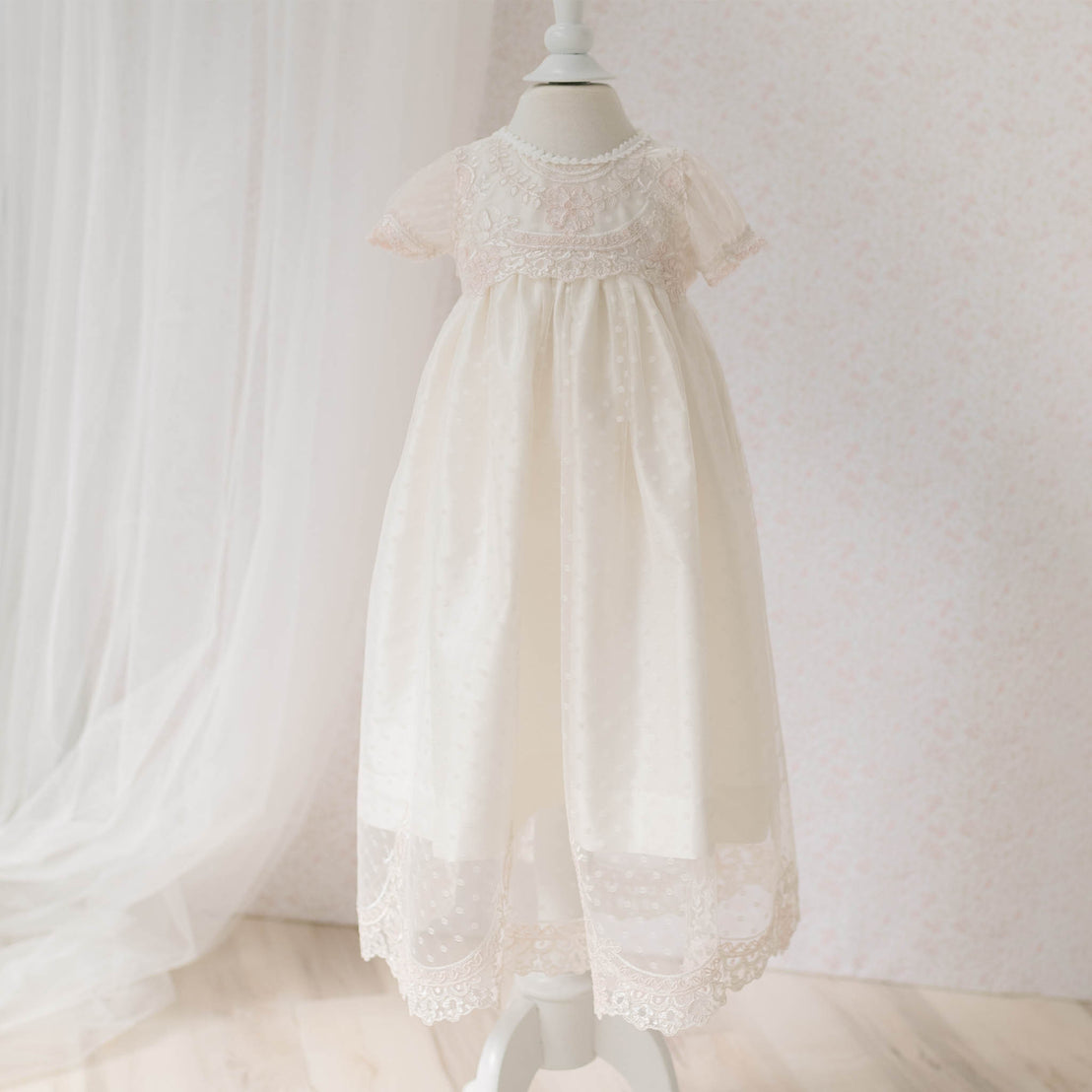 Full view of the Elizabeth Newborn Christening Dress on a mannequin against a white background and sheer lace curtain. Part of the Baby Beau & Belle Christening Dress Baby Girl Collection.