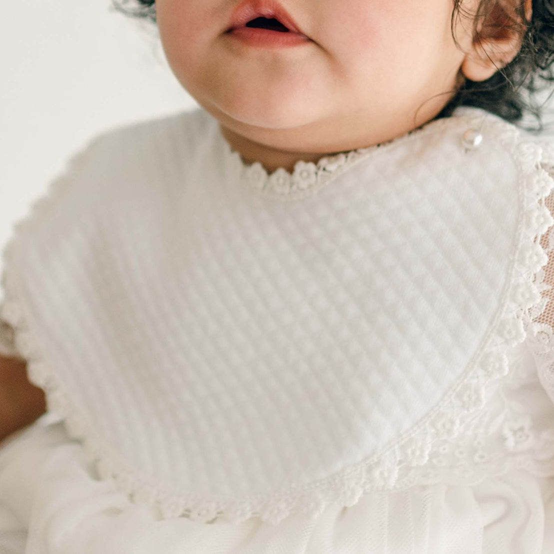 A baby girl with curly hair and expressive eyes wears the Ella Bib and Romper Dress, looking slightly off-camera.