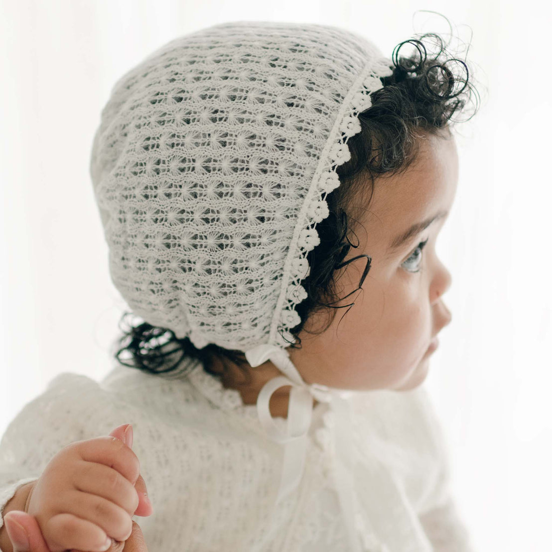 A small child with curly dark hair is wearing a crocheted white bonnet and an Eliza Christening Knit Sweater with lace detailing. The child is holding someone's hand and is turned slightly to the side, looking to the right. The background is light and blurred, adding a soft touch to the Christening Day accessory.