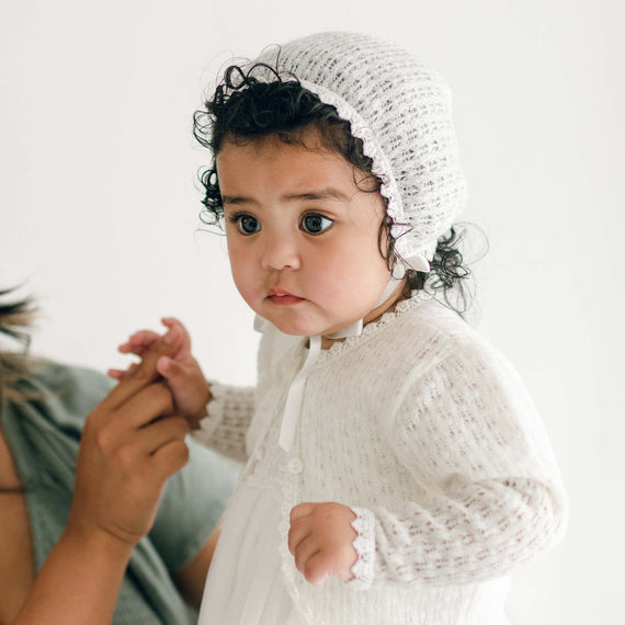 A young child with curly hair is wearing a handmade ivory knit bonnet and an Eliza Christening Knit Sweater. The child is being held by an adult who is partially visible on the left side of the image, holding the child's hand. The background is plain white, evoking a serene atmosphere suitable for a Christening Day accessory.