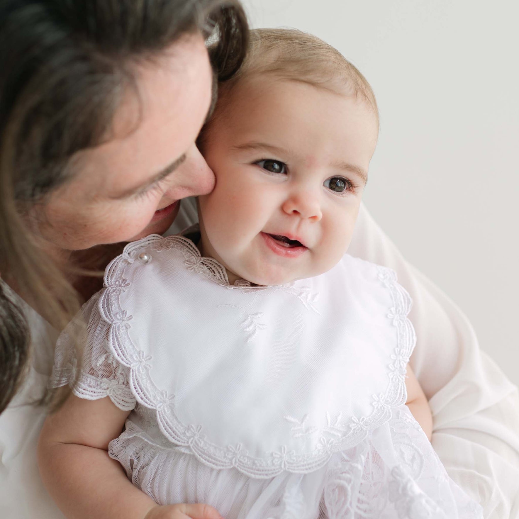 Woman holding a baby dressed in a white bib with lace details.