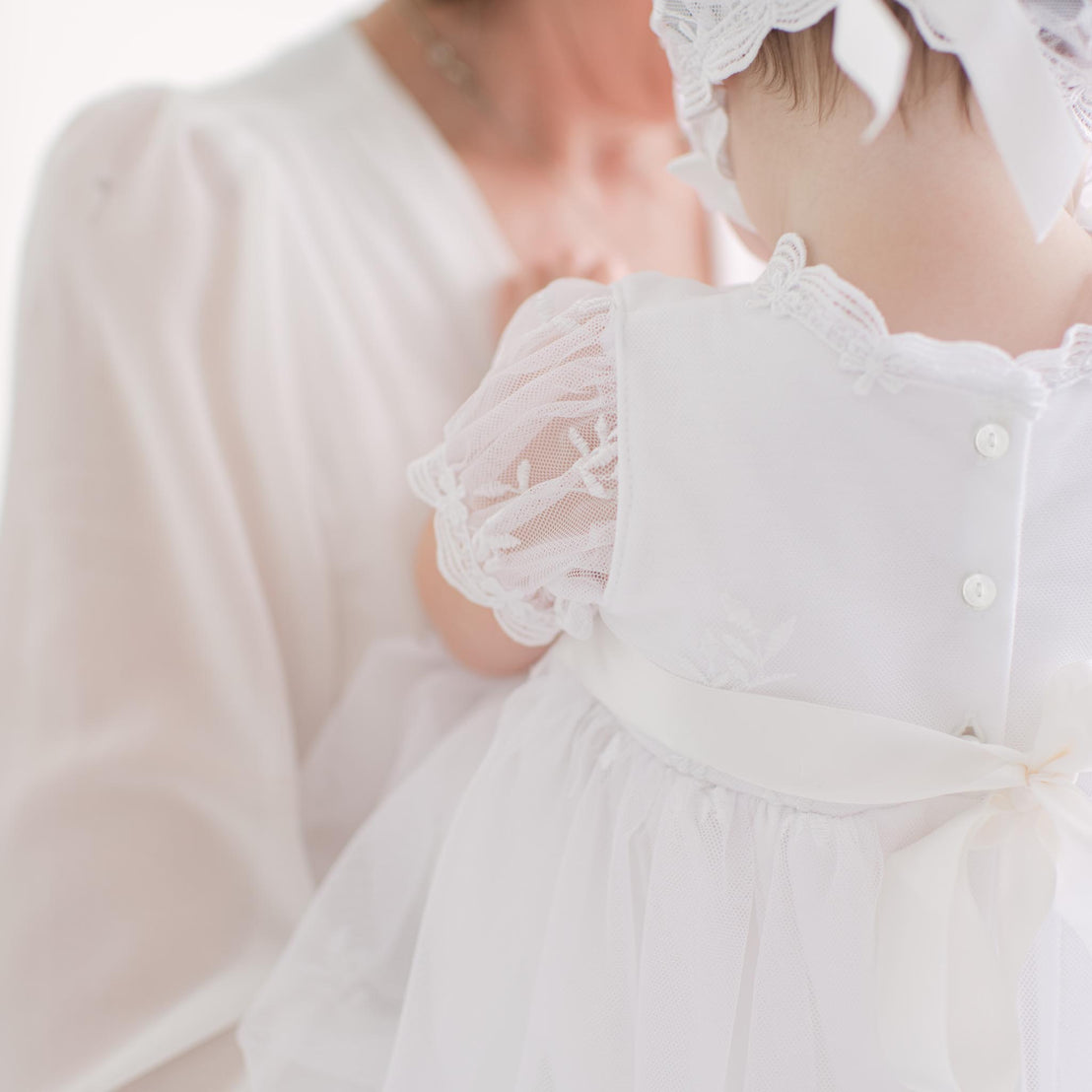 Close-up of a baby in a white outfit with lace details, held by an adult.