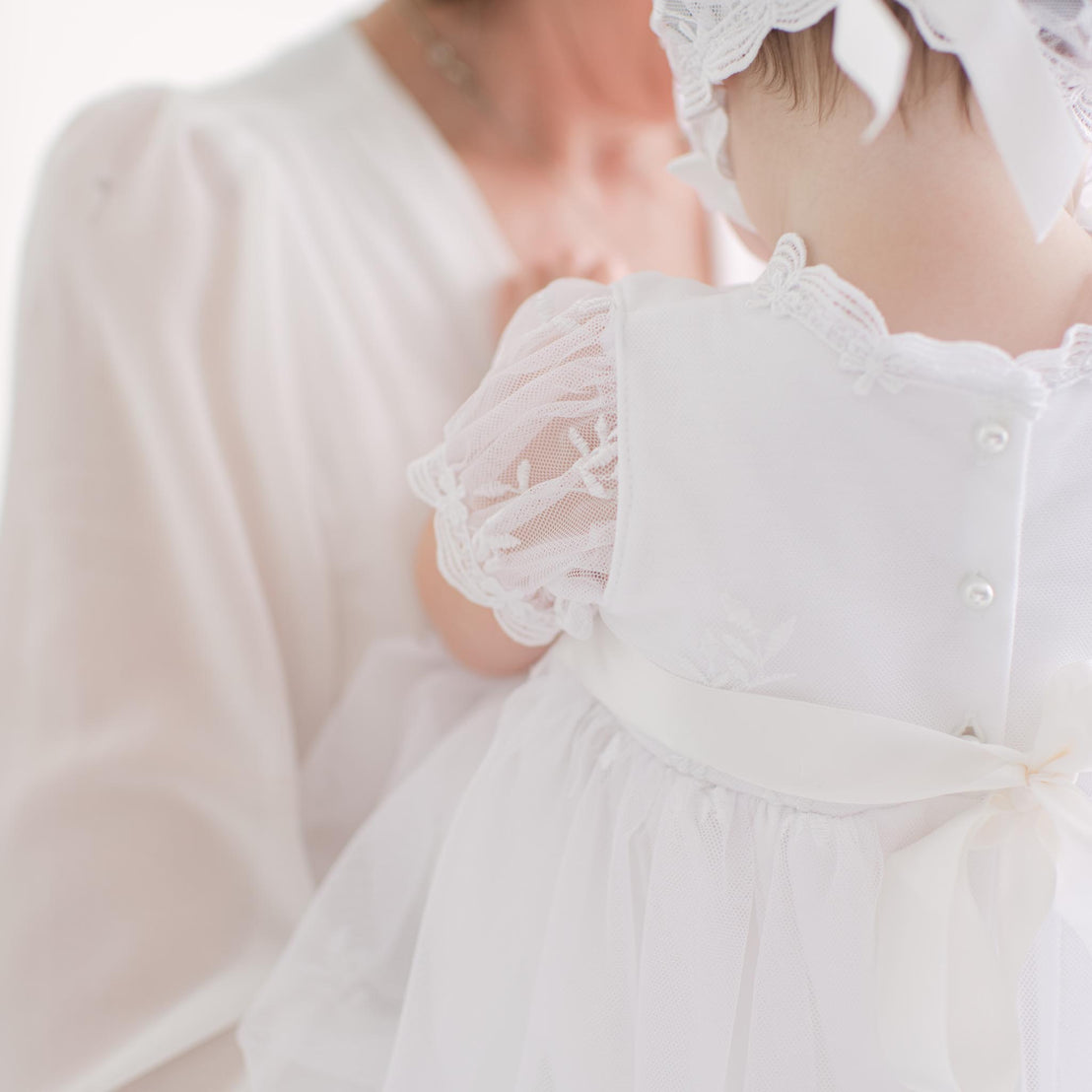 Close-up of a baby in a white outfit with lace and button details, held by an adult