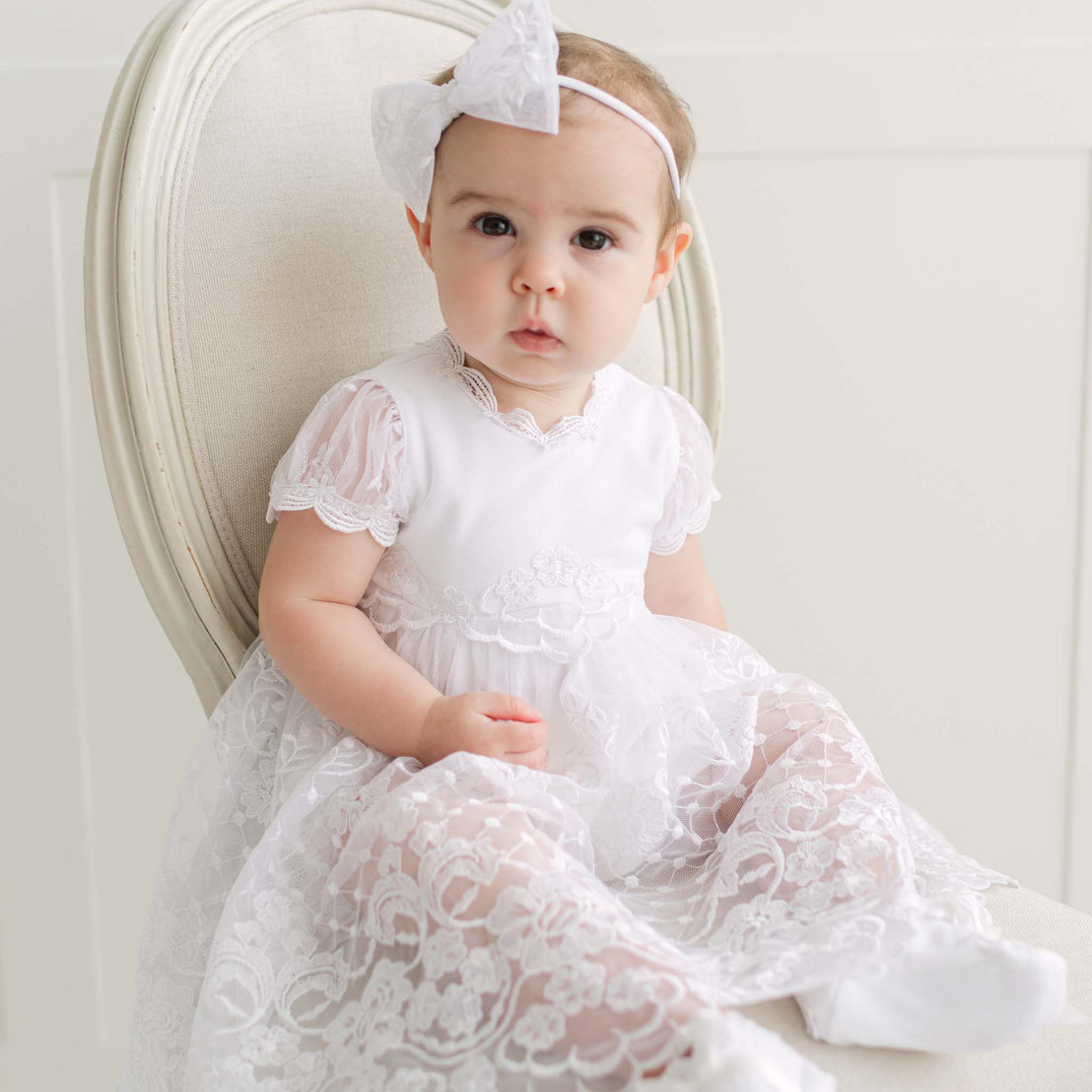 Baby in a white lace dress with a bow headband sits on a beige chair