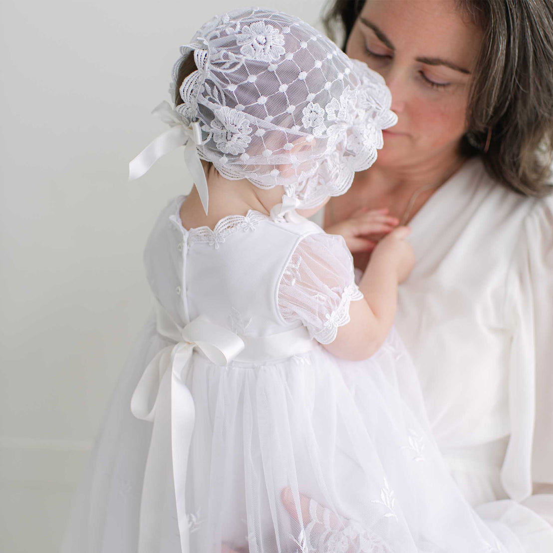 Woman holding a baby dressed in white lace dress and bonnet against a plain background
