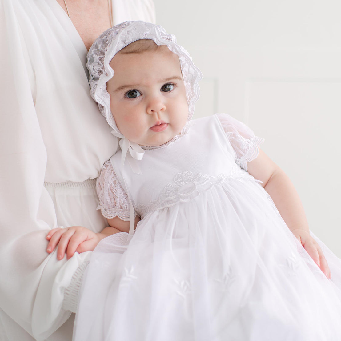 Baby in a white gown and bonnet being held by an adult against a white background
