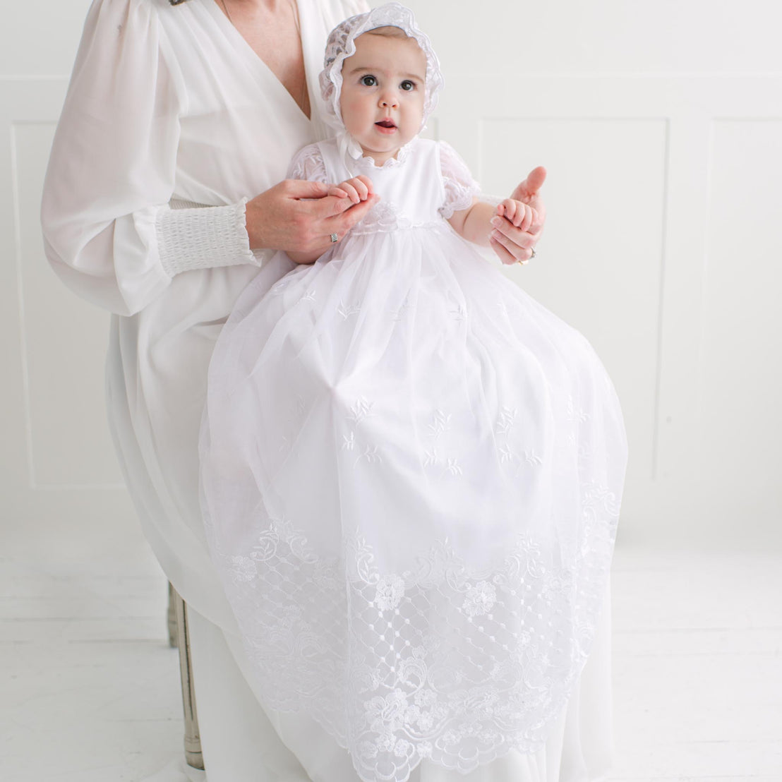Baby in a white lace gown with a bonnet, sitting on a woman's lap