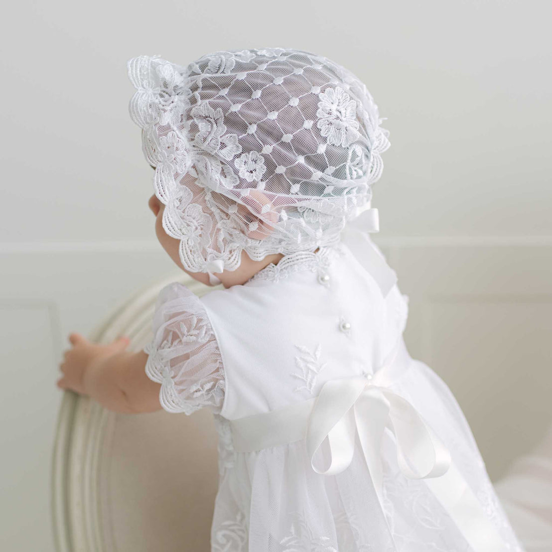 Baby wearing a white lace dress and bonnet with lace and button details against a plain background