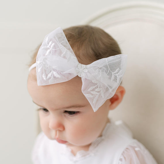 Baby wearing a white lace bow headband against a neutral background.