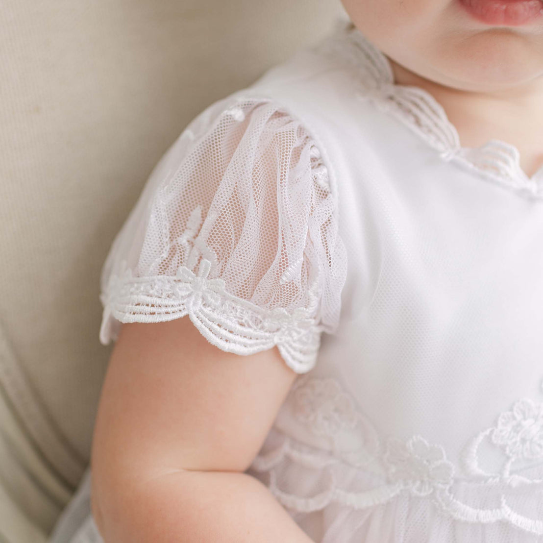 Close-up of a baby wearing a white lace dress with a beige chair in the background.