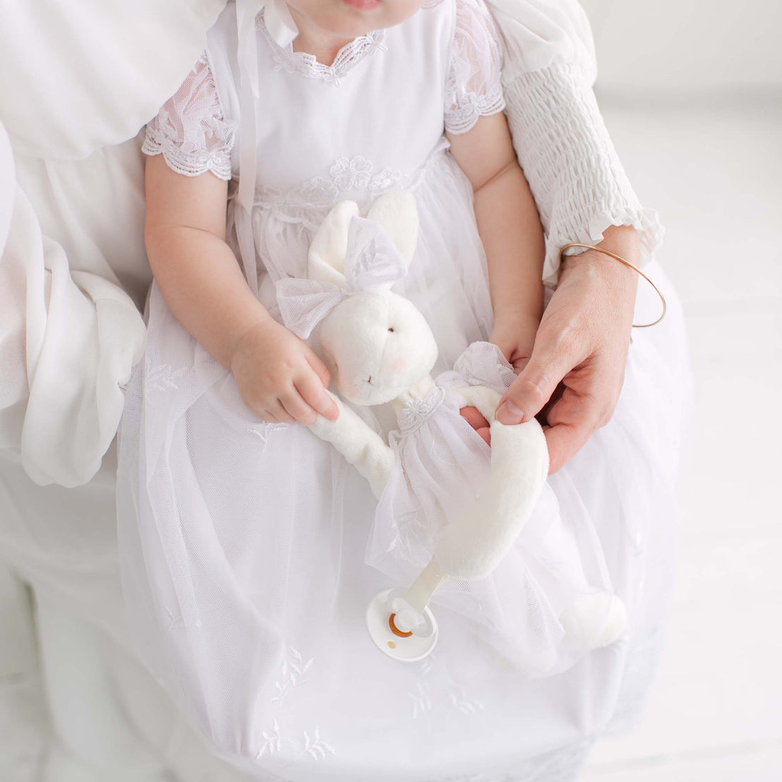 Baby in a white lace gown holding a white plush bunny toy against a white background.
