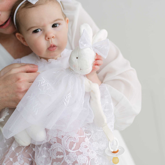 Baby in a white lace dress holding a white bunny toy with a plain background
