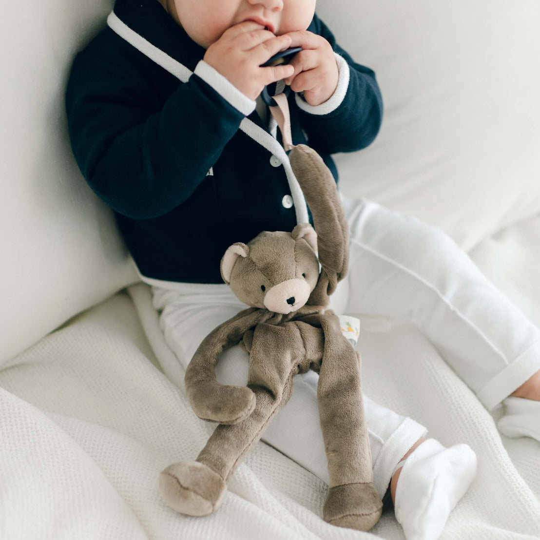 Baby boy holding on to a pacifier that is attached to the hand clip of the Elliott Bear Buddy, a floppy stuffed animal bear.