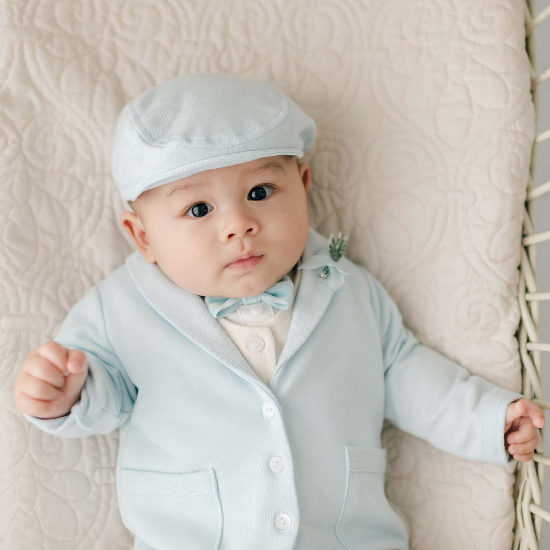 A baby, dressed in an Ezra Powder Blue 4-Piece Suit made of soft French Terry Cotton, lies on a quilted surface. The outfit includes a matching hat, jacket, and bow tie. The baby gazes directly at the camera with a neutral expression.