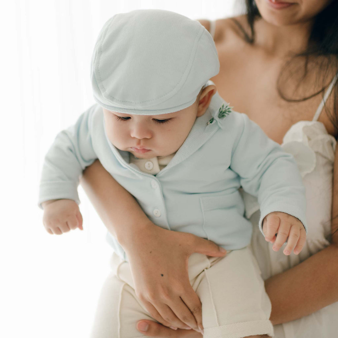 A woman holding a baby dressed in an Ezra Powder Blue 4-Piece Suit made of soft French Terry Cotton. The baby, looking down in the light blue outfit and matching hat, is gently supported by the woman's arms. The background is softly lit, creating a serene atmosphere.