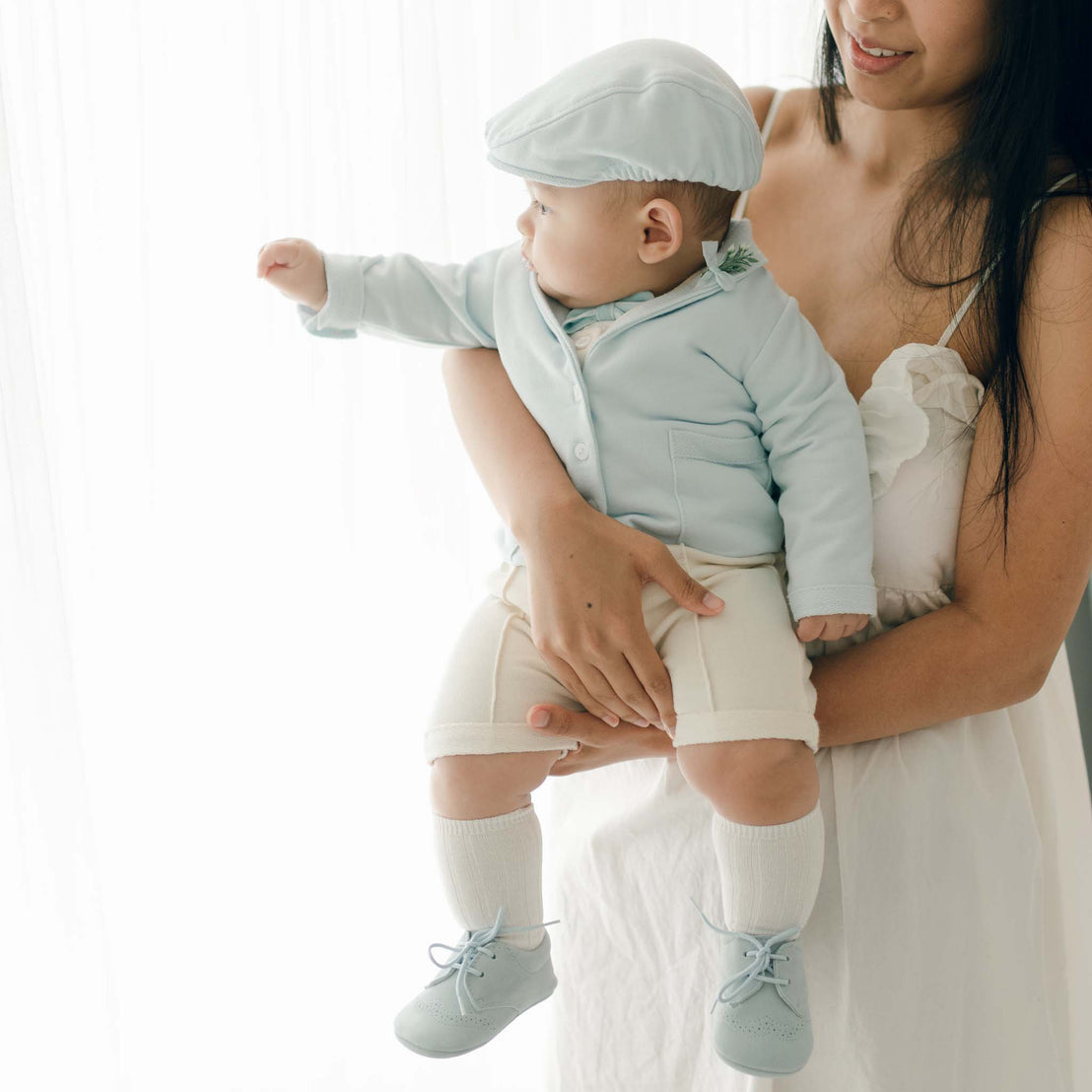 A baby wearing a light blue cap, jacket, and shoes with white shorts and knee-high socks is being held by a woman in a sleeveless white dress. The baby, dressed in an Ezra Powder Blue 4-Piece Suit made from French Terry Cotton, is looking towards the light source coming from a window.