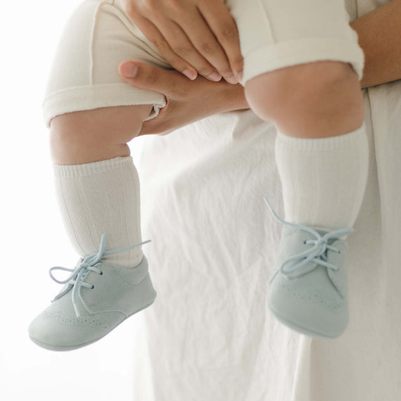 A person is holding a baby dressed in white clothing, with Cream Ribbed Socks and pale blue lace-up shoes. The focus is on the baby's legs and feet, adorned in ribbed pattern attire. The background is softly lit, creating a calm atmosphere that feels like a cozy treat.