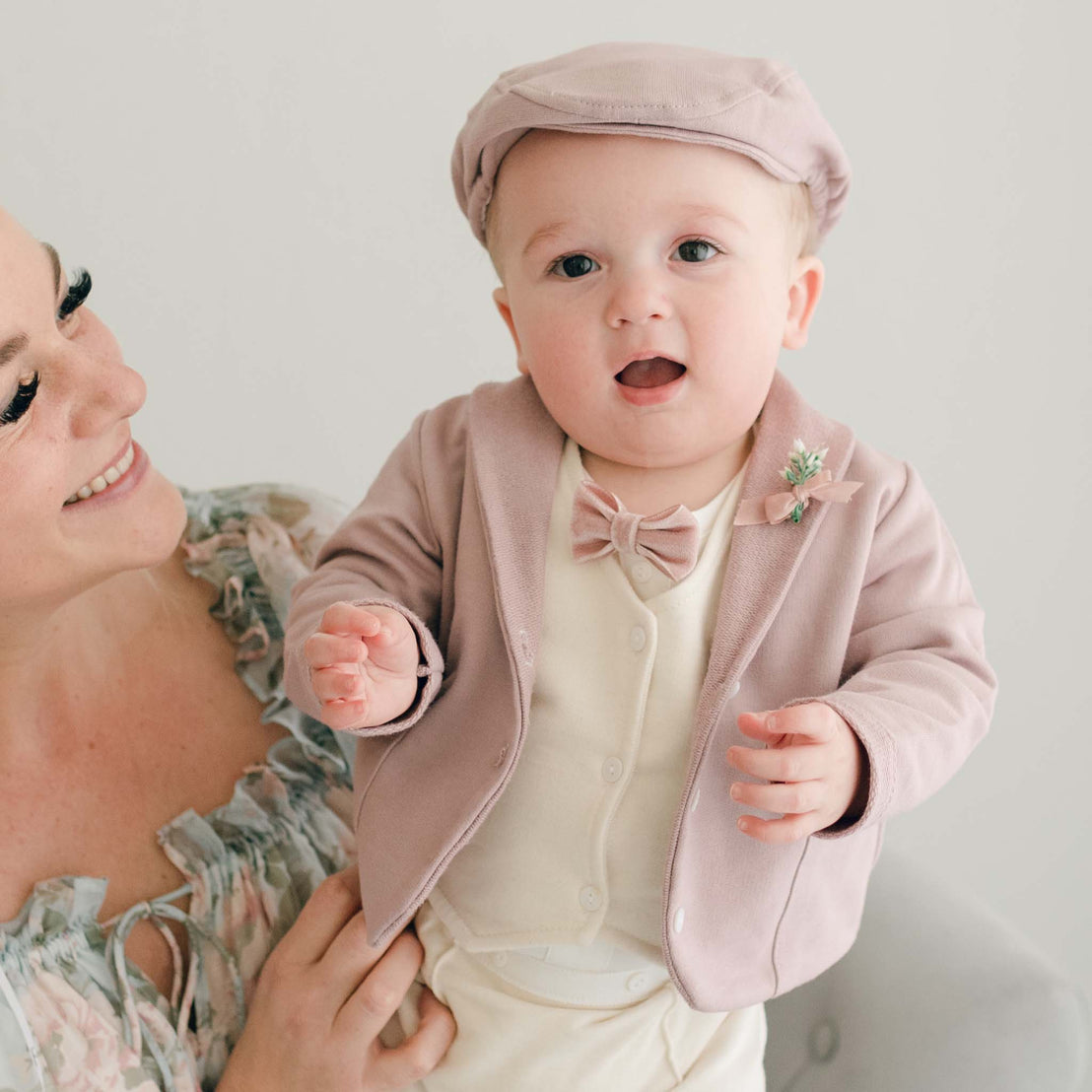 Closer detail of baby boy standing his mother's lap. He is wearing the Ezra 4-Piece Mauve Suit with matching Newsboy Cap, mauve velvet bow tie and boutonniere
