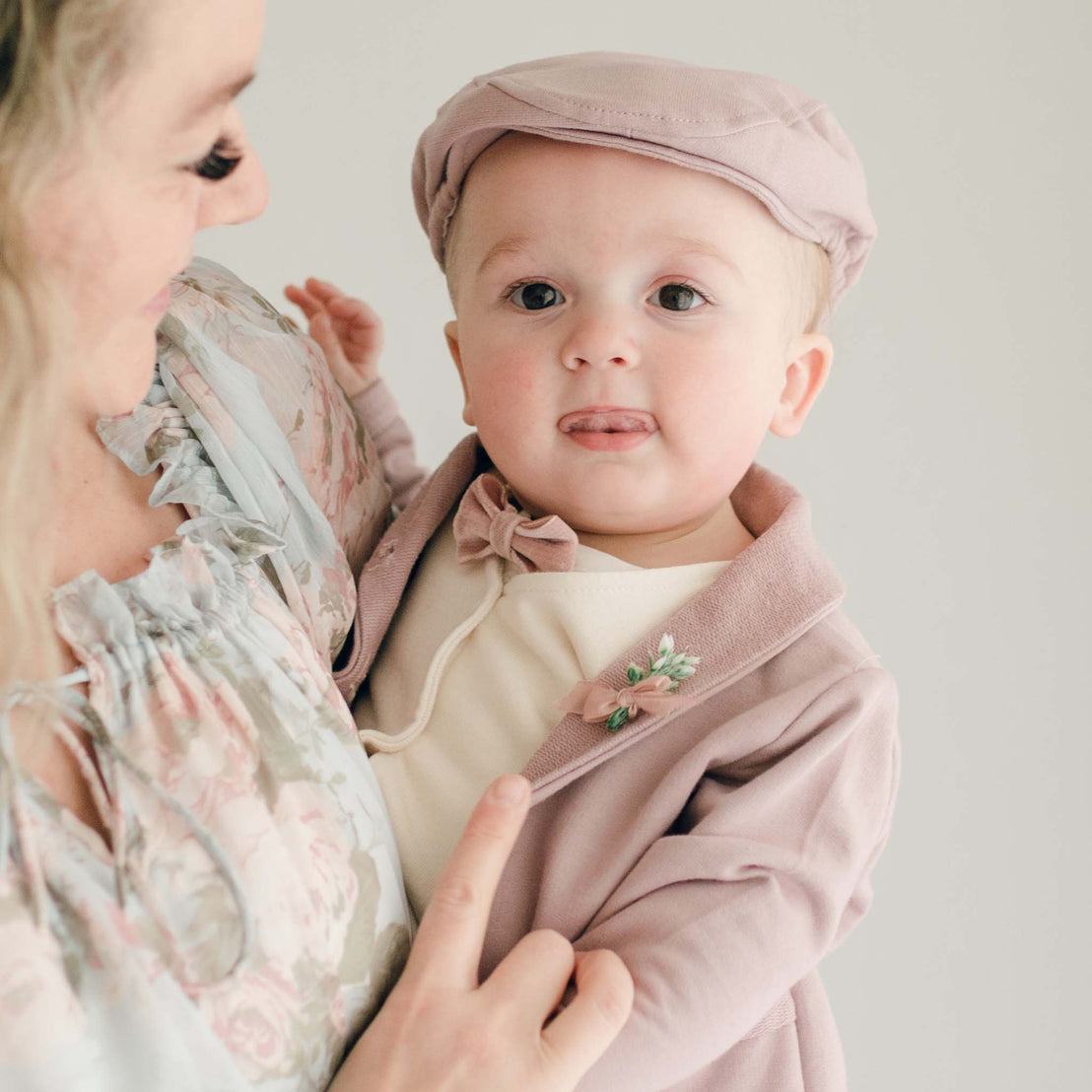 Mother holding her baby boy. The boy is wearing Baby boy standing his mother's lap. He is wearing the Ezra 4-Piece Mauve Suit with matching Newsboy Cap, mauve velvet bow tie and boutonniere